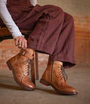 A person wearing Bed Stu's Endure rugged lace-up ankle boots with leather lining, brown pants, and a white long-sleeve top sits on a wooden chair against a brick wall, showcasing effortless rugged sophistication.