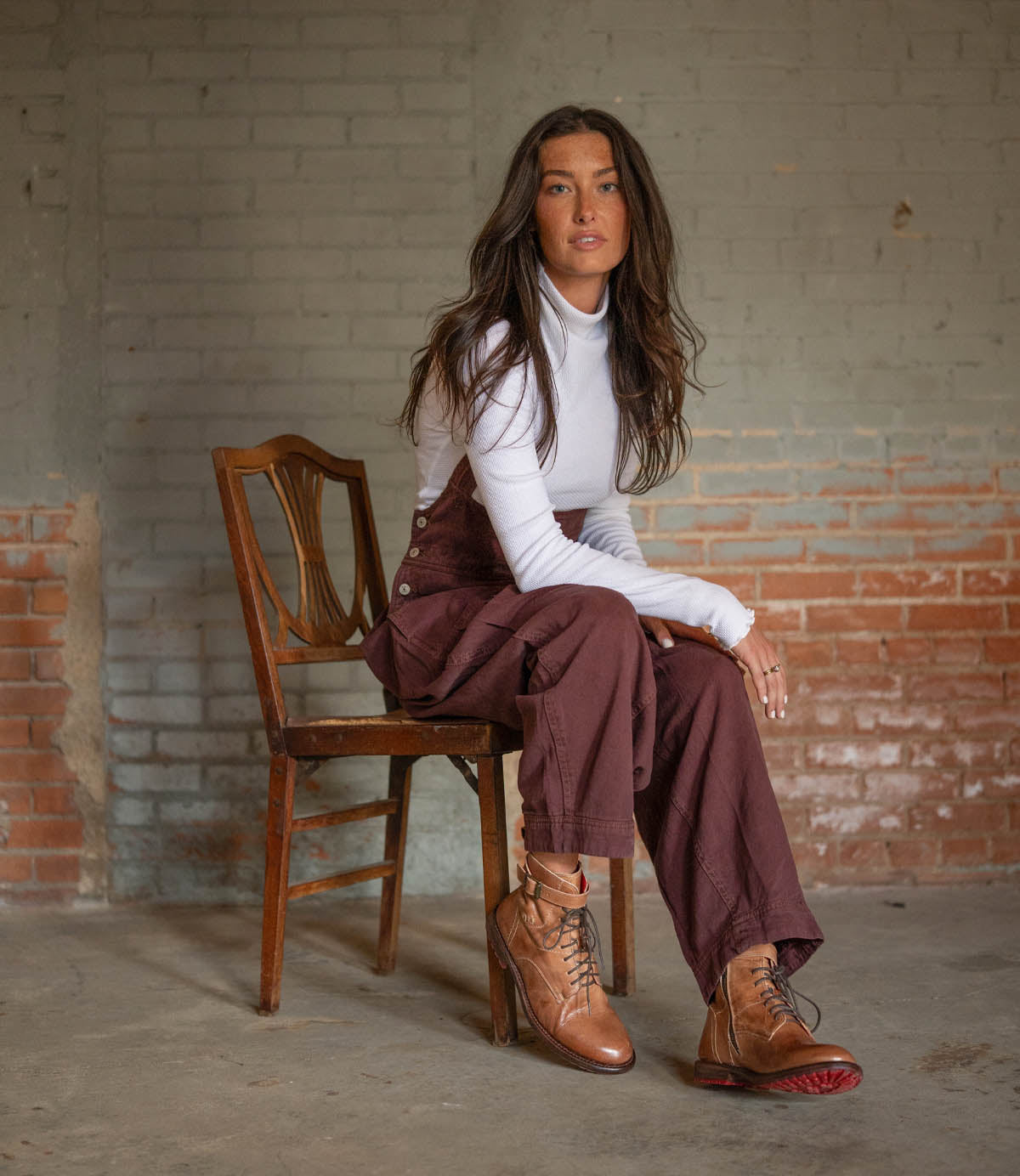 A woman with long brown hair in a white turtleneck, maroon overalls, and Bed Stu Endure lace-up ankle boots sits on a wooden chair against brick walls and concrete floor, embodying rugged sophistication.