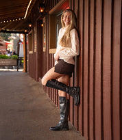 A woman with long hair smiles as she leans against a wooden wall outside, wearing Bed Stu's Essence black knee-high riding boots styled with a cream sweater and brown skirt.