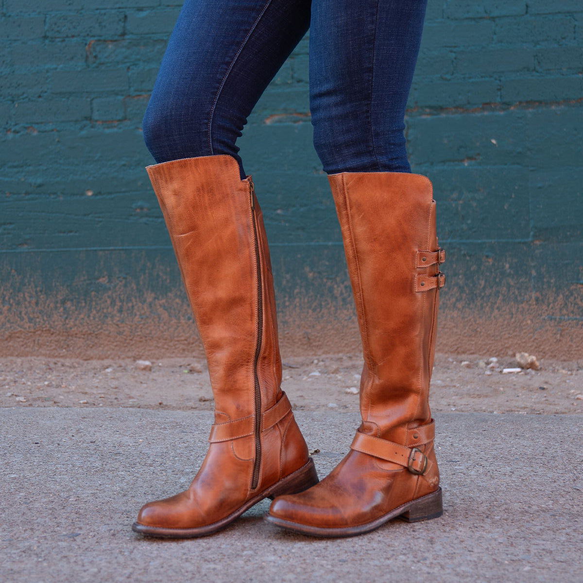 A person in Bed Stu Essence hand-finished brown leather knee-high riding boots and dark blue jeans stands on a concrete surface before a teal brick wall.