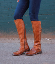 A person in Bed Stu Essence hand-finished brown leather knee-high riding boots and dark blue jeans stands on a concrete surface before a teal brick wall.