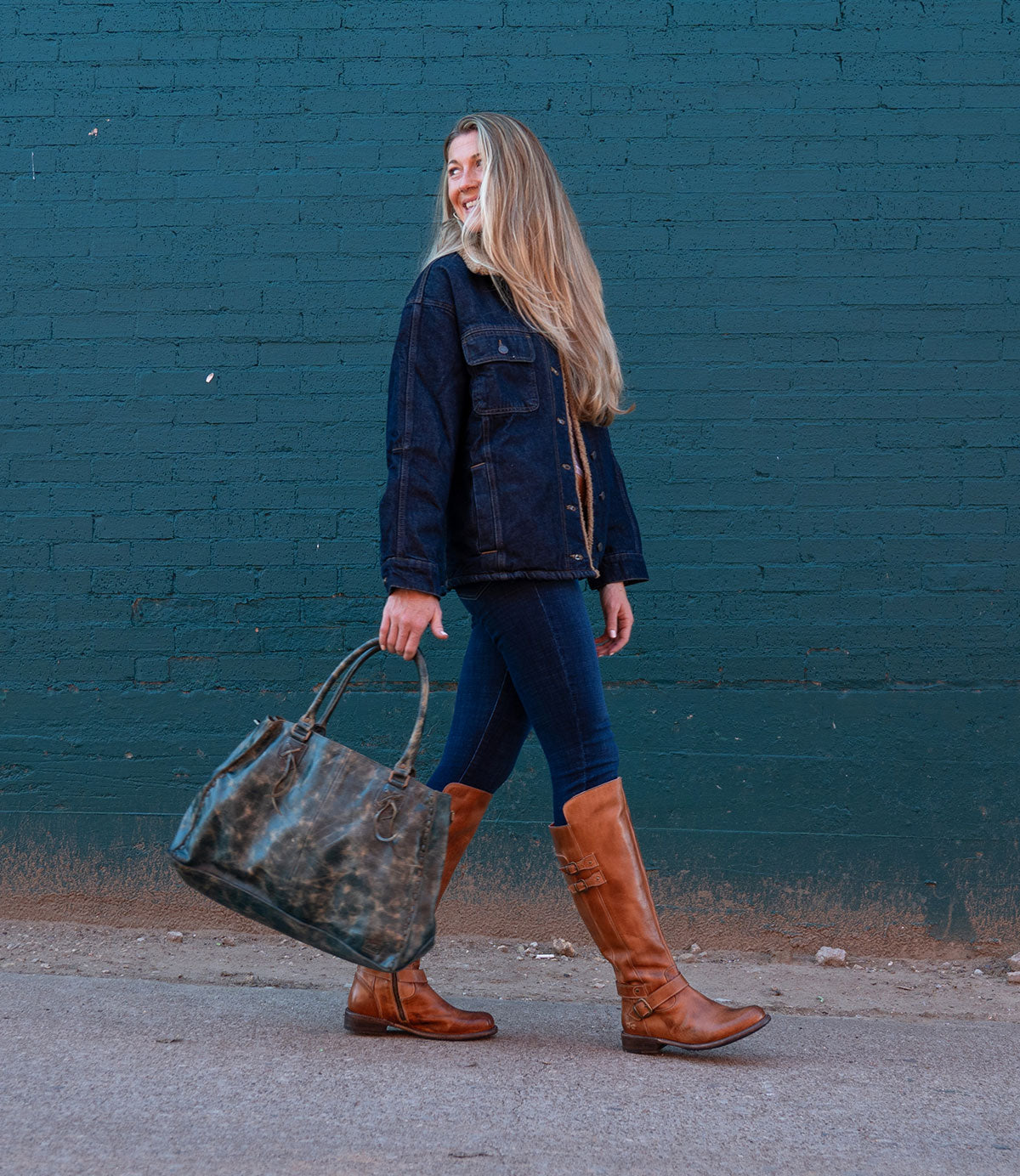 A woman with long blonde hair walks by a teal brick wall, wearing a dark jacket, jeans, and Bed Stu’s Essence knee-high riding boots with equestrian design, carrying a large dark green bag.