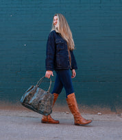A woman with long blonde hair walks by a teal brick wall, wearing a dark jacket, jeans, and Bed Stu’s Essence knee-high riding boots with equestrian design, carrying a large dark green bag.