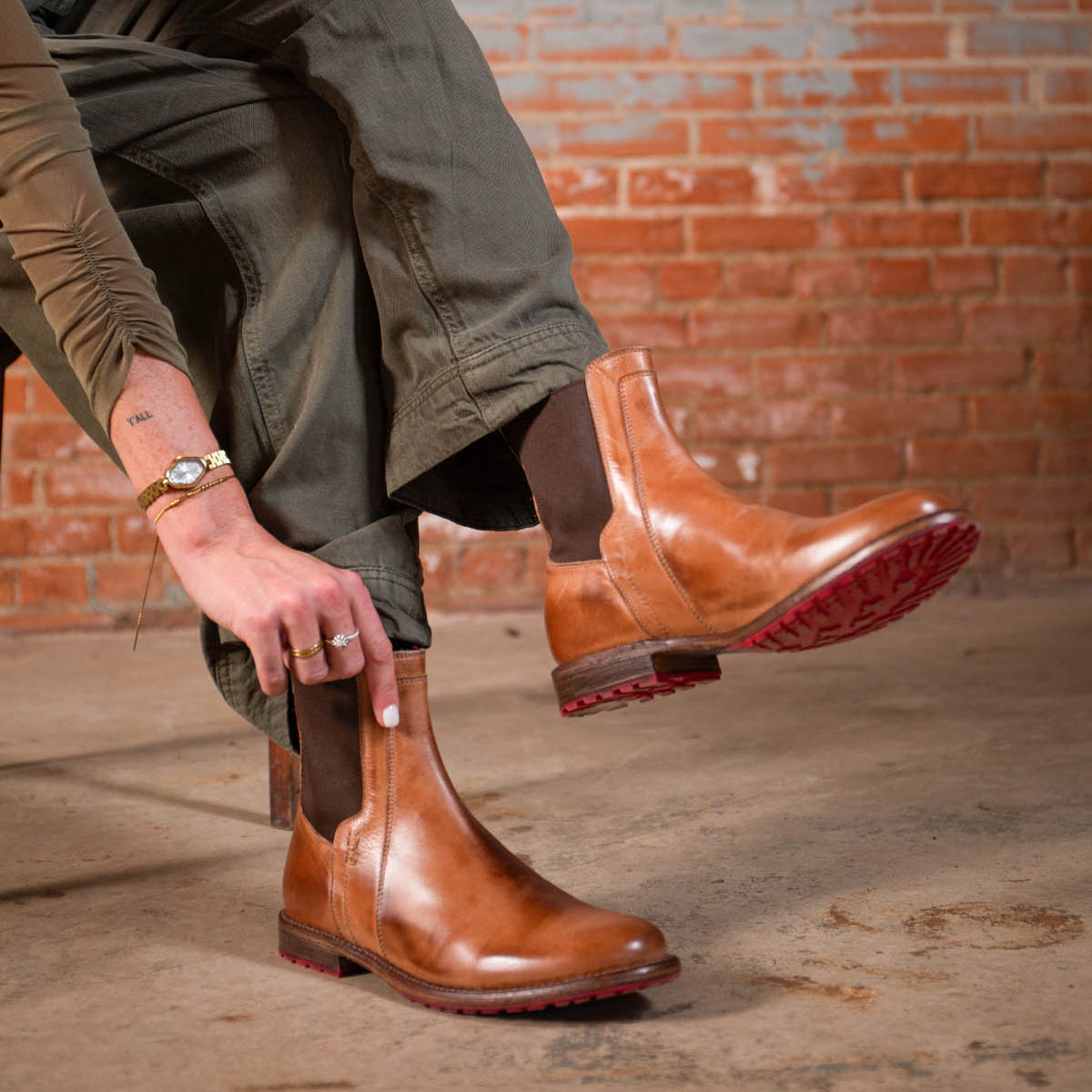 A person sits on a bench against a brick wall, wearing olive pants and putting on Bed Stu Ethic brown leather Chelsea boots with Vibram soles and red accents.
