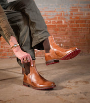 A person sits on a bench against a brick wall, wearing olive pants and putting on Bed Stu Ethic brown leather Chelsea boots with Vibram soles and red accents.