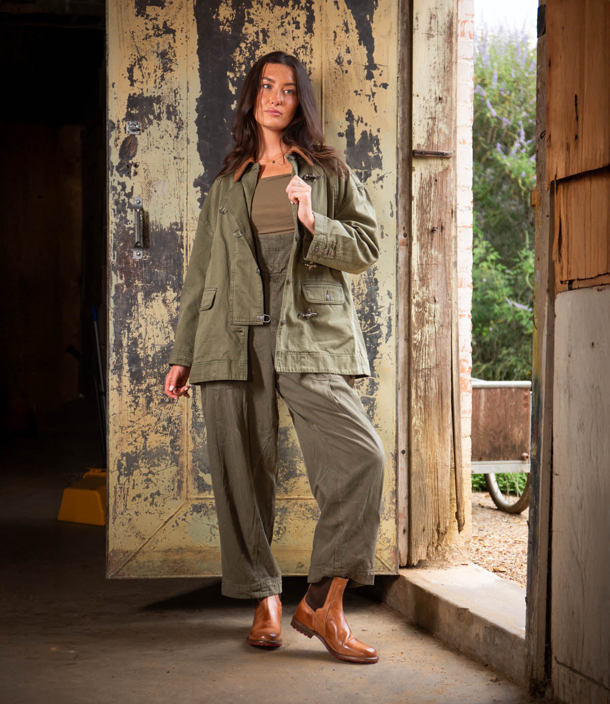 A woman stands in a doorway wearing Bed Stu Ethic vegetable-tanned leather Chelsea boots, paired with an olive green jacket, matching pants, and a brown top against rustic, weathered surroundings.