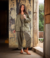 A woman stands in a doorway wearing Bed Stu Ethic vegetable-tanned leather Chelsea boots, paired with an olive green jacket, matching pants, and a brown top against rustic, weathered surroundings.