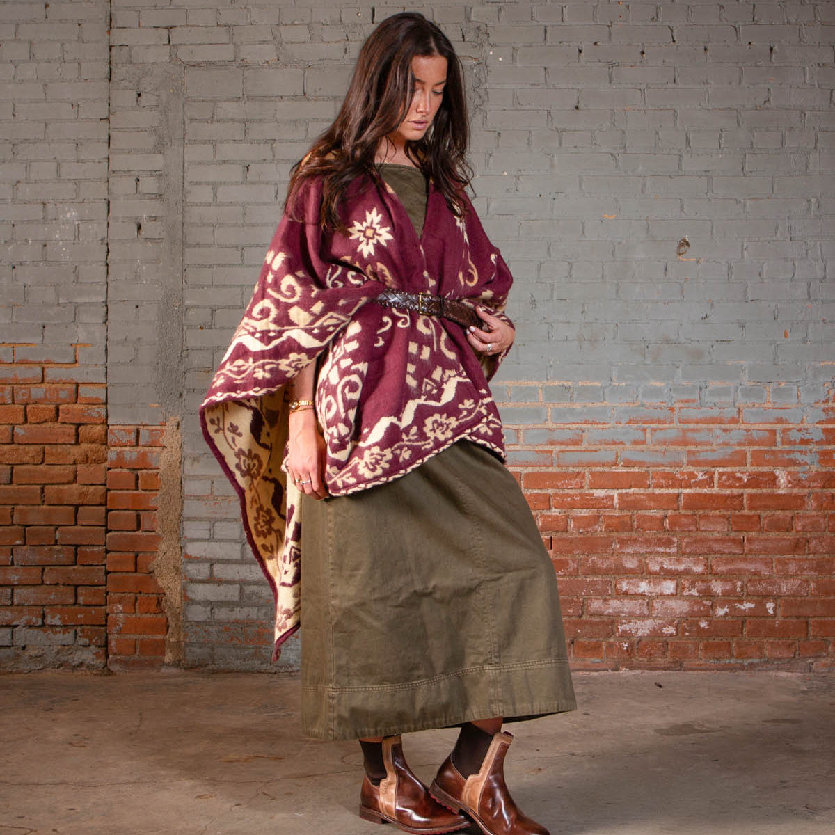A woman stands indoors against a painted brick wall, wearing the Bed Stu Ethic Chelsea boots with a rugged Vibram sole, styled with a maroon patterned shawl, olive dress, and brown belt.