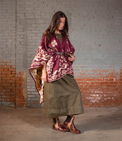 A woman stands indoors against a painted brick wall, wearing the Bed Stu Ethic Chelsea boots with a rugged Vibram sole, styled with a maroon patterned shawl, olive dress, and brown belt.