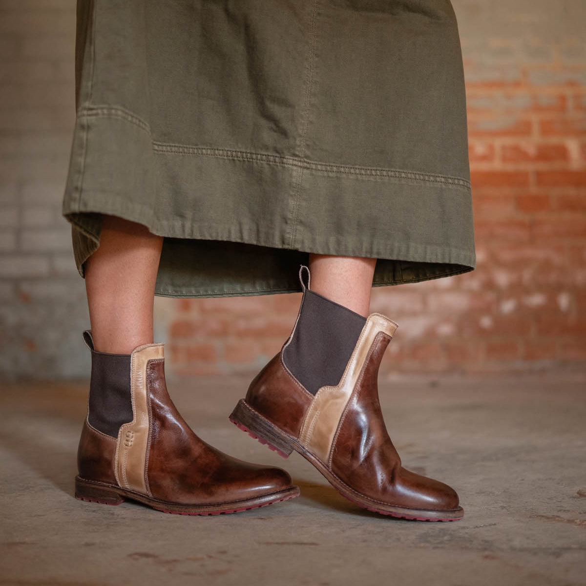 Someone wearing a long olive-green skirt and Bed Stu's Ethic brown vegetable-tanned leather Chelsea boots stands on a concrete floor against a brick wall.