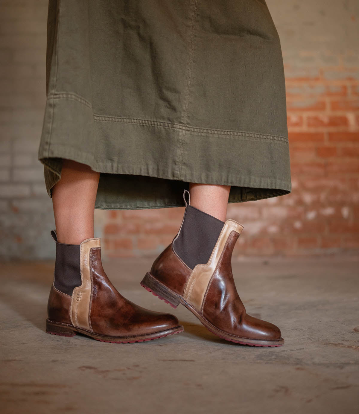 Someone wearing a long olive-green skirt and Bed Stu's Ethic brown vegetable-tanned leather Chelsea boots stands on a concrete floor against a brick wall.