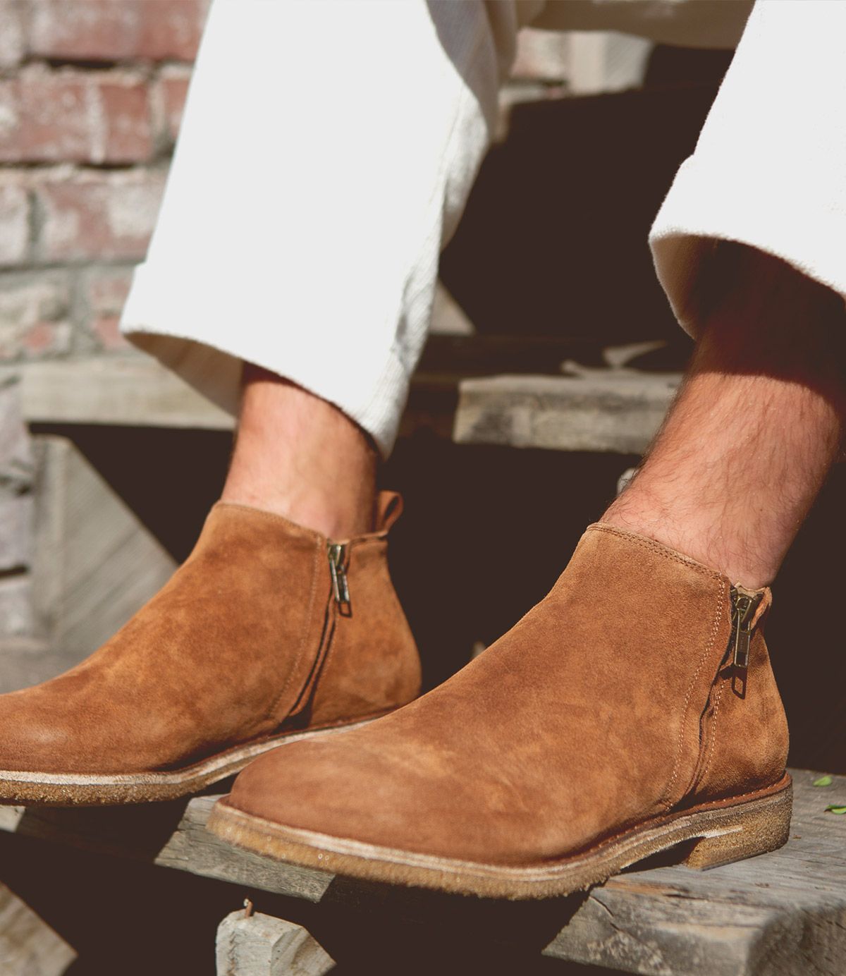 A person wearing light-colored pants and Bed Stu Evade handcrafted brown suede ankle boots with dual zippers, seated on wooden steps in an outdoor setting.