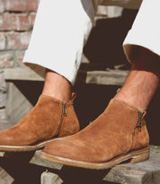 A person wearing light-colored pants and Bed Stu Evade handcrafted brown suede ankle boots with dual zippers, seated on wooden steps in an outdoor setting.
