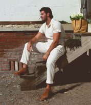 A man wearing a white shirt and pants sits on outdoor wooden steps beside a brick wall. A woven basket with green plants is placed on the steps next to him, subtly highlighting his premium Bed Stu Evade suede shoes handcrafted in Italy.