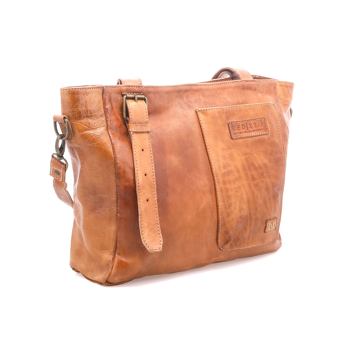 The Bed Stu Explicit LTC is a light brown seasoned leather bag with an adjustable strap, front pocket, visible stitching, and rectangular embossing. This utility bag has a rustic appeal and is shown against a white background.