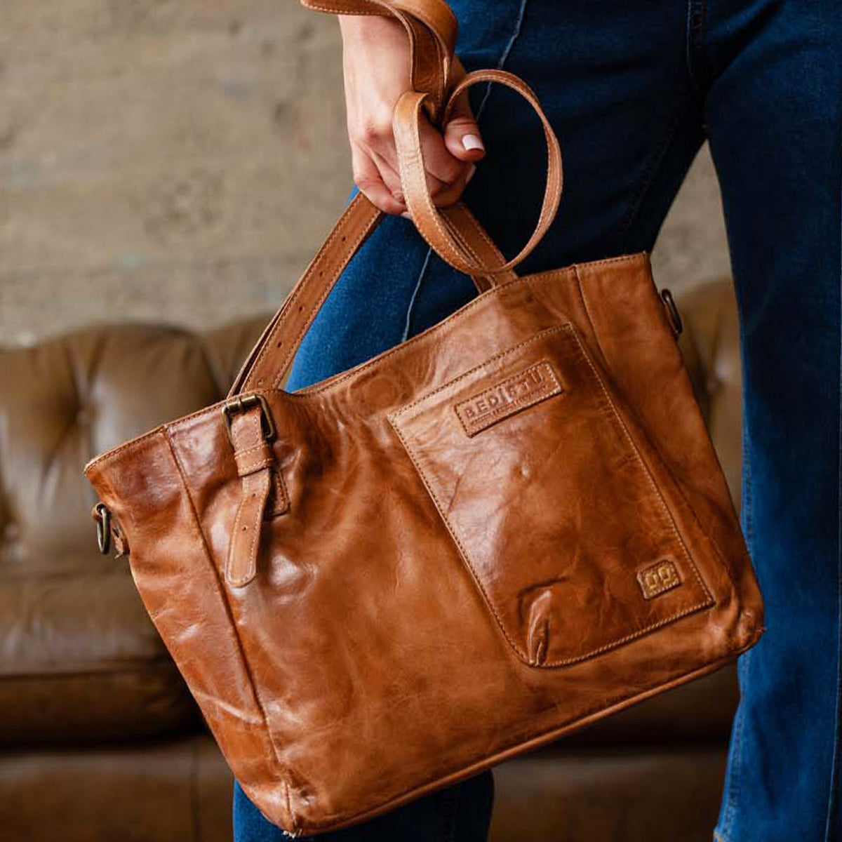 A person holding the Explicit LTC leather utility bag by Bed|Stü, featuring a front pocket and buckle detail, stands in front of a brown couch.