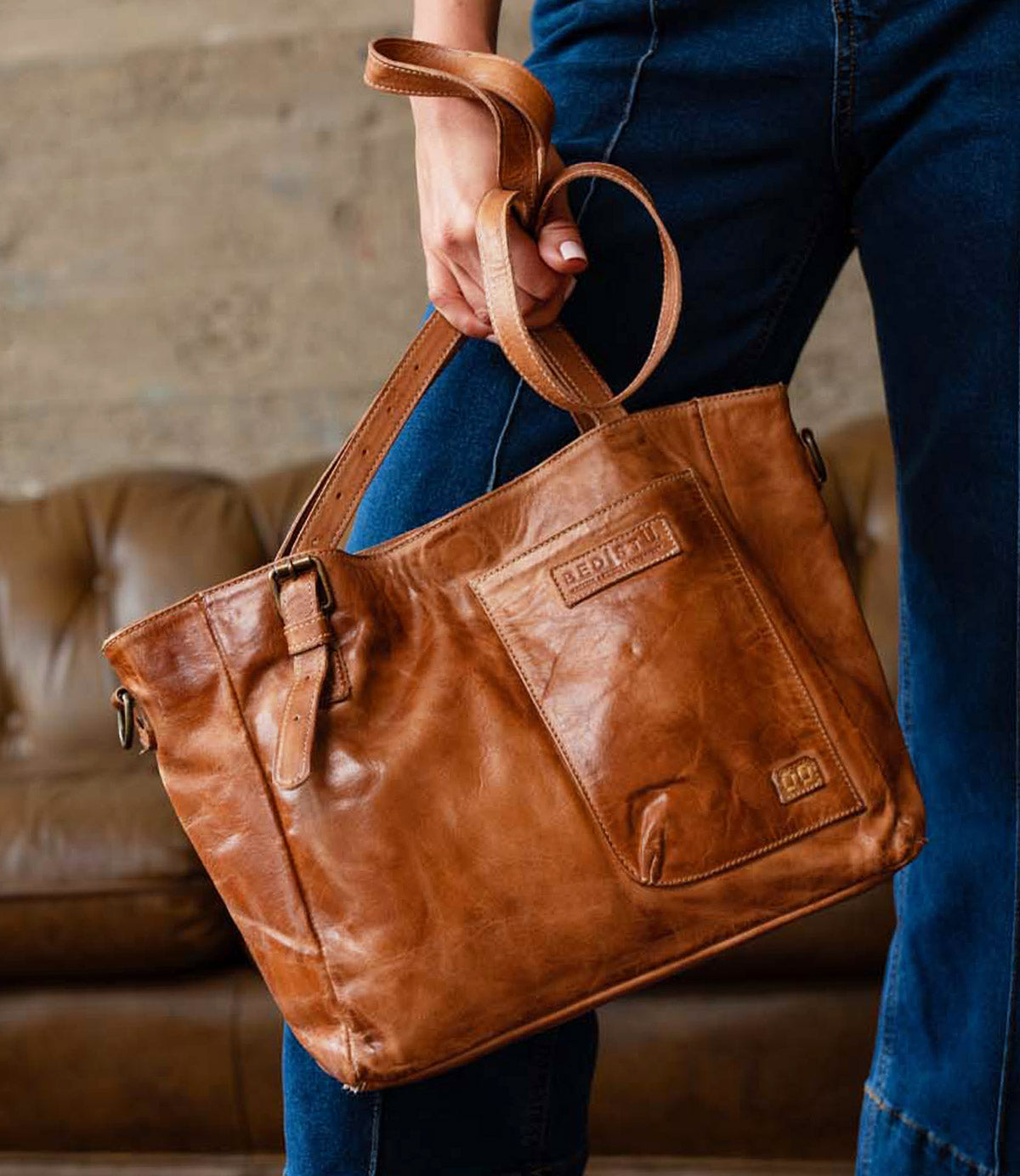 A person holding the Explicit LTC leather utility bag by Bed|Stü, featuring a front pocket and buckle detail, stands in front of a brown couch.