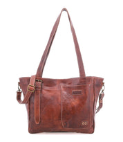 The Bed Stu Explicit LTC is a brown leather shoulder bag with two handles, an adjustable strap, visible stitching, a front pocket, and a small rectangular logo patch for rustic appeal and versatile utility.
