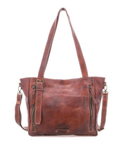 The Bed Stu Explicit LTC is a rustic brown leather tote bag with two top handles, front zippered pockets, and a detachable shoulder strap, shown against a white background.