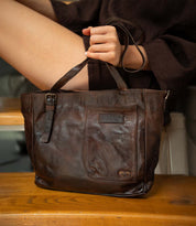 A person sitting on stairs holds the Bed|Stü Explicit LTC, a seasoned leather bag in dark brown featuring two handles and an external pocket, highlighting the utility craftsmanship and rustic appeal of this accessory.