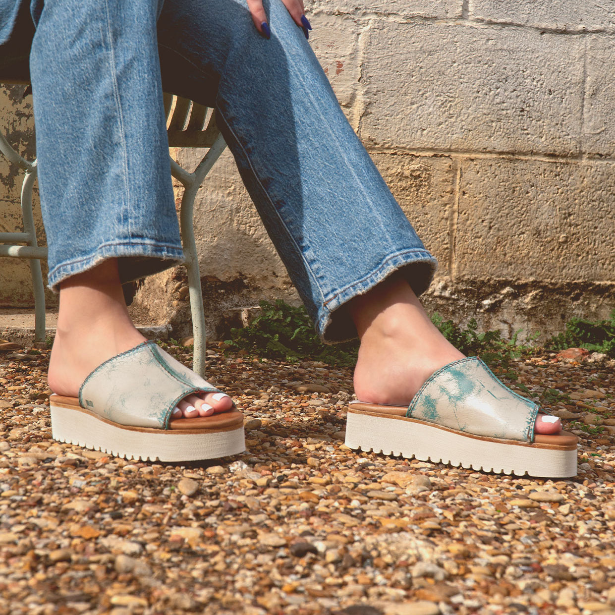 A person sits on a metal chair wearing Bed Stu's eco-friendly Fairlee II sandals with blue jeans, feet resting on a gravel surface beside a textured wall.