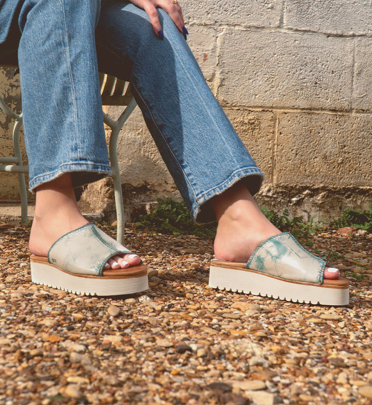 A person sits on a metal chair wearing Bed Stu's eco-friendly Fairlee II sandals with blue jeans, feet resting on a gravel surface beside a textured wall.