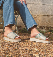A person sits on a metal chair wearing Bed Stu's eco-friendly Fairlee II sandals with blue jeans, feet resting on a gravel surface beside a textured wall.