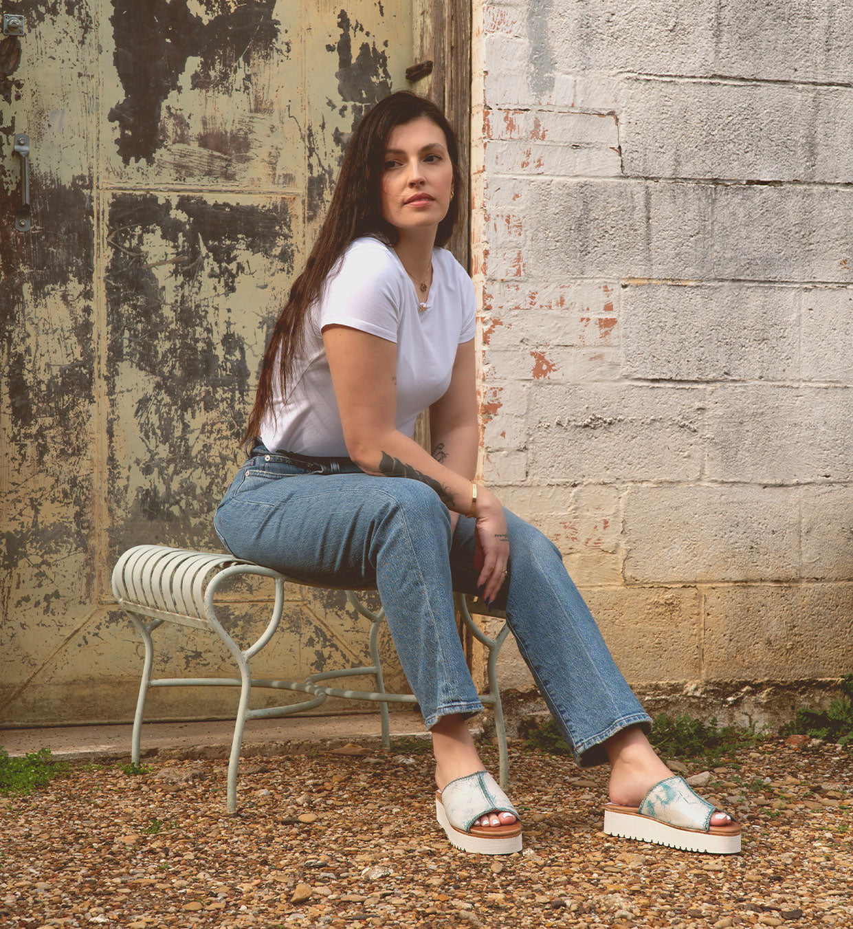 A woman wearing the Fairlee II flatform leather slide sandals by Bed Stu sits on a metal bench outdoors in a white t-shirt and jeans, with a distressed wall and door in the background.
