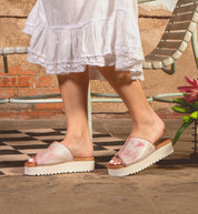 A person stands on a checkered floor near a metal chair and pink flower, wearing Bed Stu Fairlee II pink platform slide sandals with cushioned footbed and a white dress.