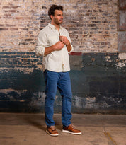 A man stands indoors by a weathered brick wall, wearing a light button-up shirt, blue jeans, and Bed Stu Fairman low-top leather sneakers featuring a comfortable insole.