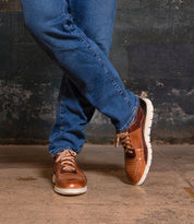 A person wears Bed Stu Fairman brown low-top leather sneakers with comfortable insoles and blue jeans, standing with legs crossed on a concrete floor against a dark, textured wall.