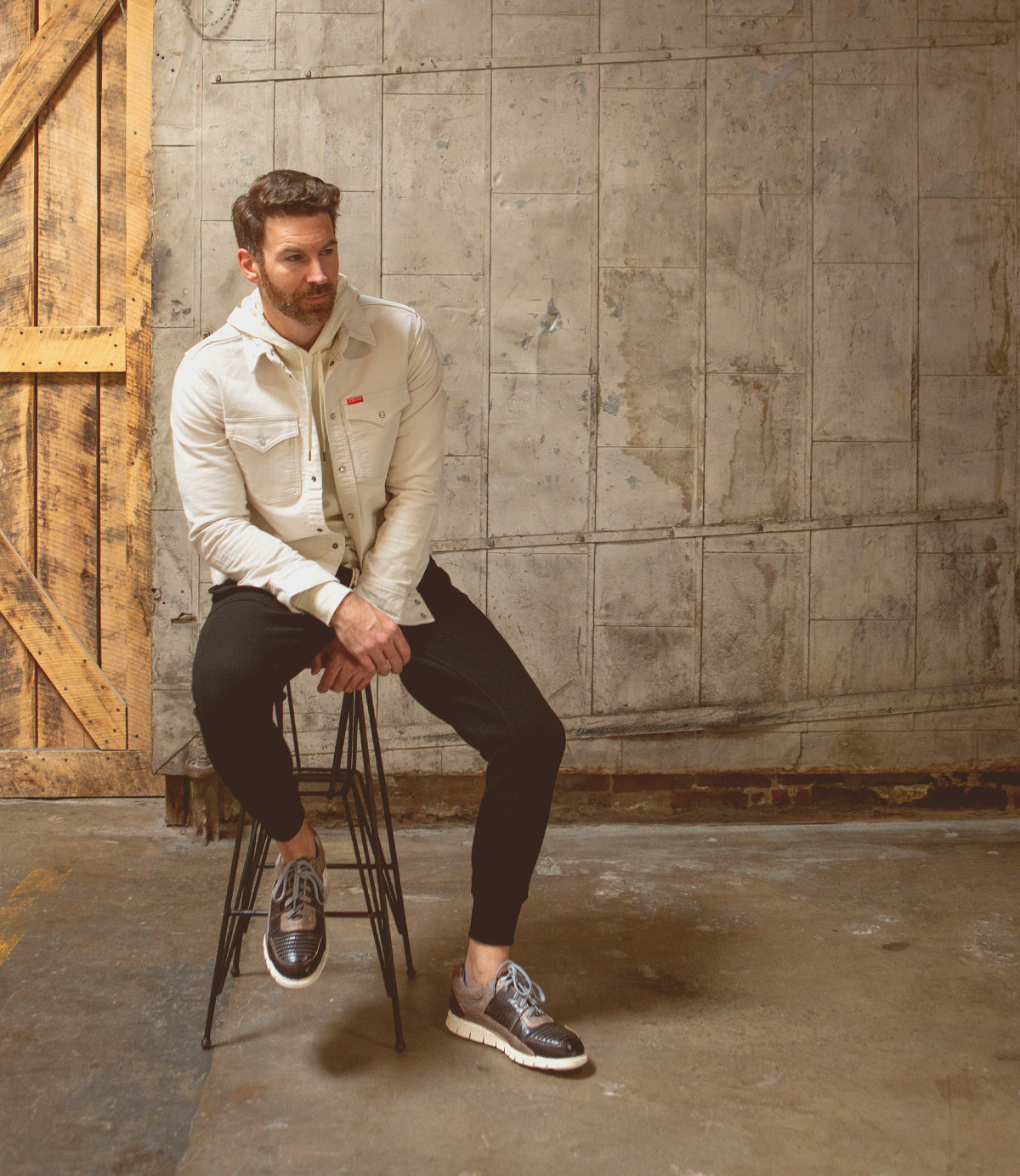 A man, with distinct character, sitting on a stool in a warehouse wearing Bed Stu low-top leather sneakers.