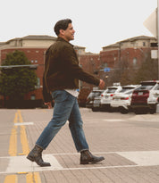 A person in a brown jacket and jeans, wearing Bed Stu's Barton leather high-top sneakers with breathable insoles, crosses a street on a crosswalk, with parked cars and brick buildings in the background.