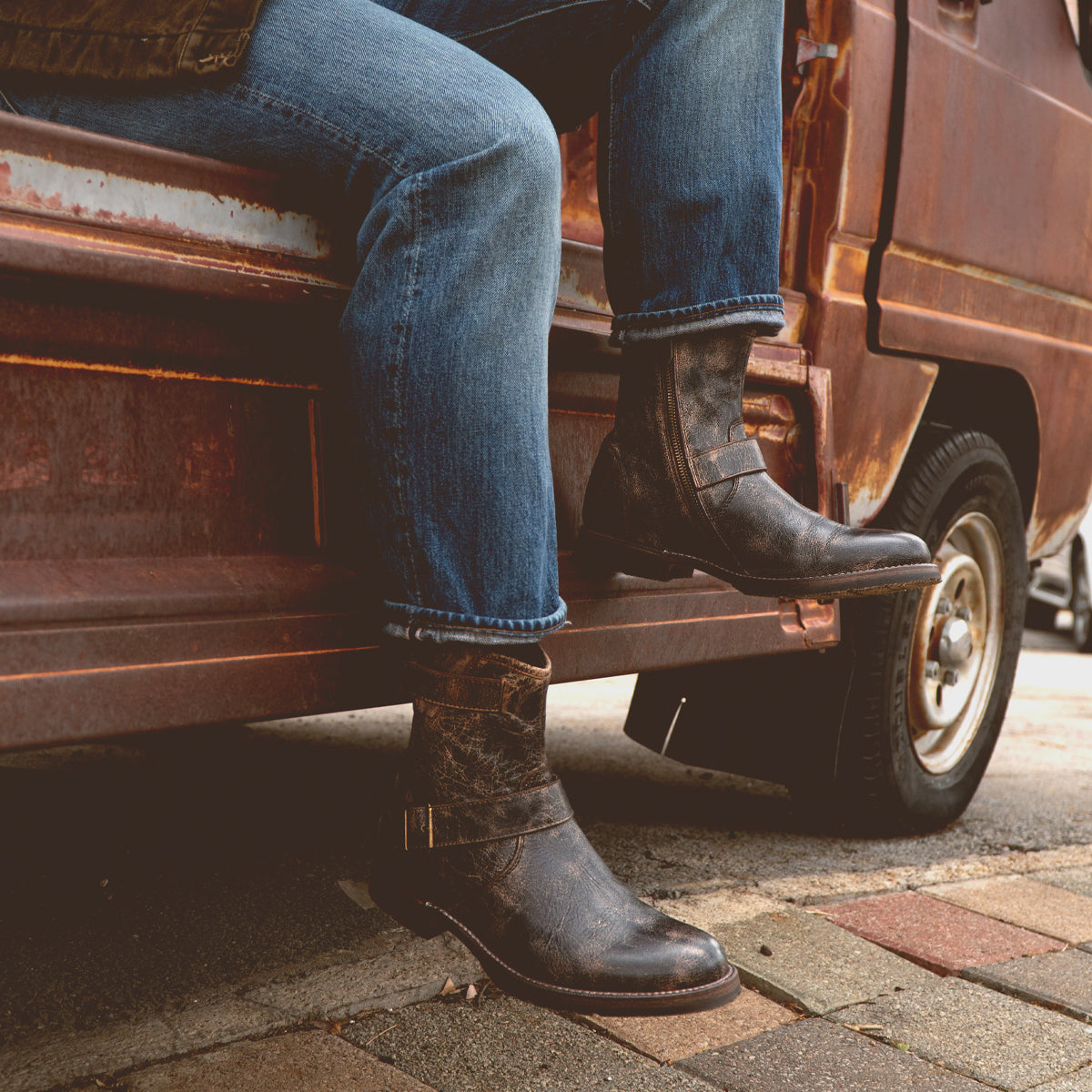 A person sits on the open door of a rusty vehicle, wearing Bed Stu's Barton sneakers featuring leather high-tops and a breathable insole, against a stone pavement backdrop.