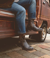A person sits on the open door of a rusty vehicle, wearing Bed Stu's Barton sneakers featuring leather high-tops and a breathable insole, against a stone pavement backdrop.