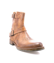 The Bed Stu Fellow is a tan moto-inspired ankle boot with two side buckles and a low heel, crafted from rugged leather, shown on a white background.