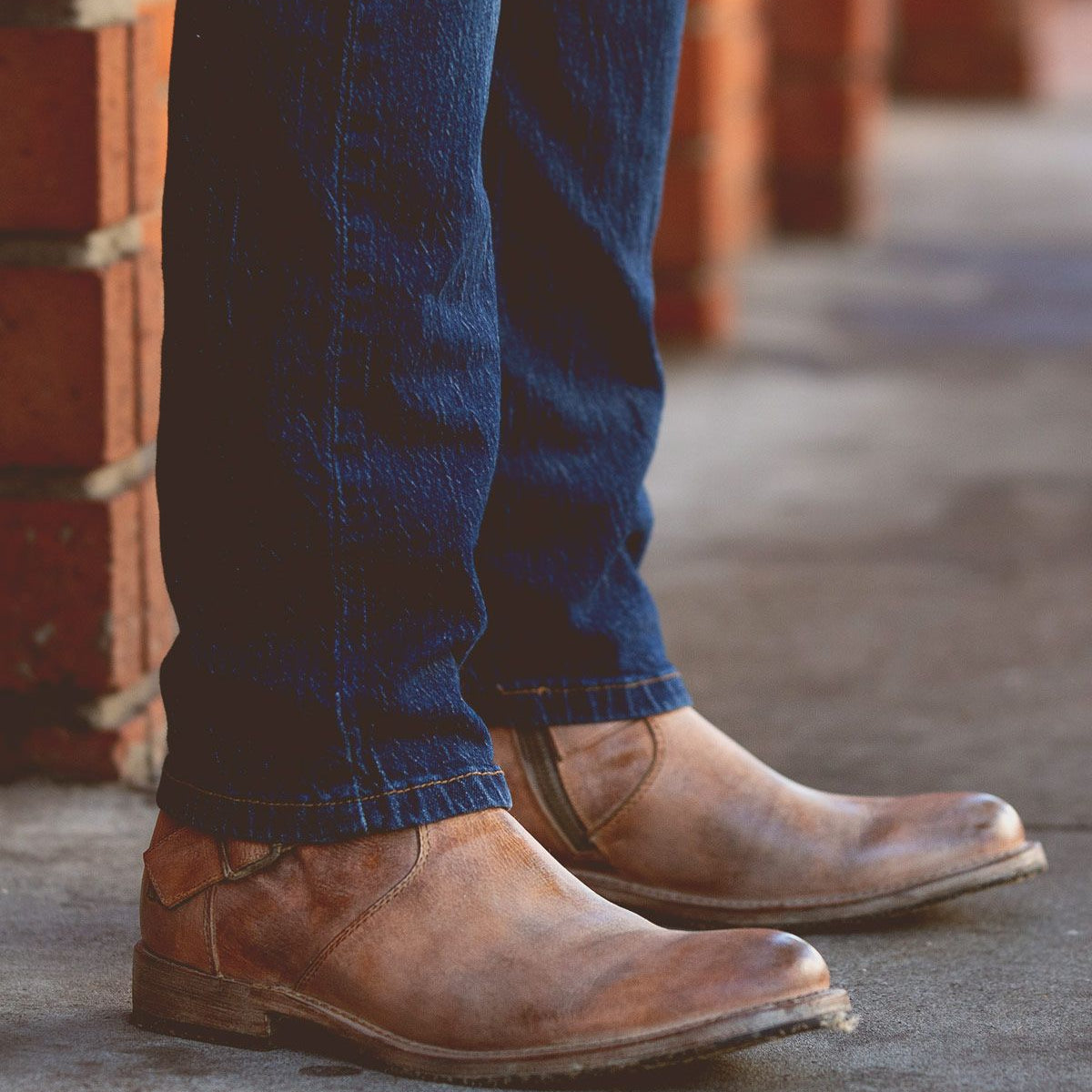 A person sporting the Fellow boots by Bed Stu in a moto-inspired leather design, paired with dark blue jeans, stands on a concrete sidewalk near red brick walls.