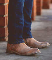 A person sporting the Fellow boots by Bed Stu in a moto-inspired leather design, paired with dark blue jeans, stands on a concrete sidewalk near red brick walls.