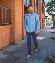 A man in a blue jacket and jeans strolls confidently along a sidewalk beside a brick building, his moto-inspired look completed with stylish Fellow leather boots by Bed Stu.