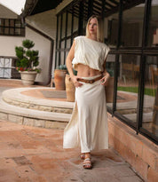 A woman stands on a tiled patio in a cream crop top and long skirt, completing her look with Bed Stu's Fibril bohemian sandals featuring an adjustable strap, near large windows and potted plants.