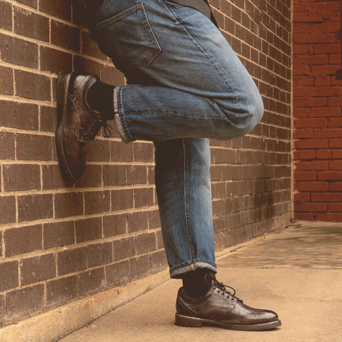 A person in jeans and Bed Stu Barton leather high-top sneakers leans against a brick wall, one leg crossed over the other.