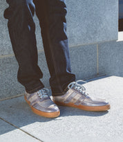 A person exudes casual cool in jeans and Bed Stu's Firepower gray men's sneakers, standing confidently on a stone surface, showcasing a classic vulcanized style.