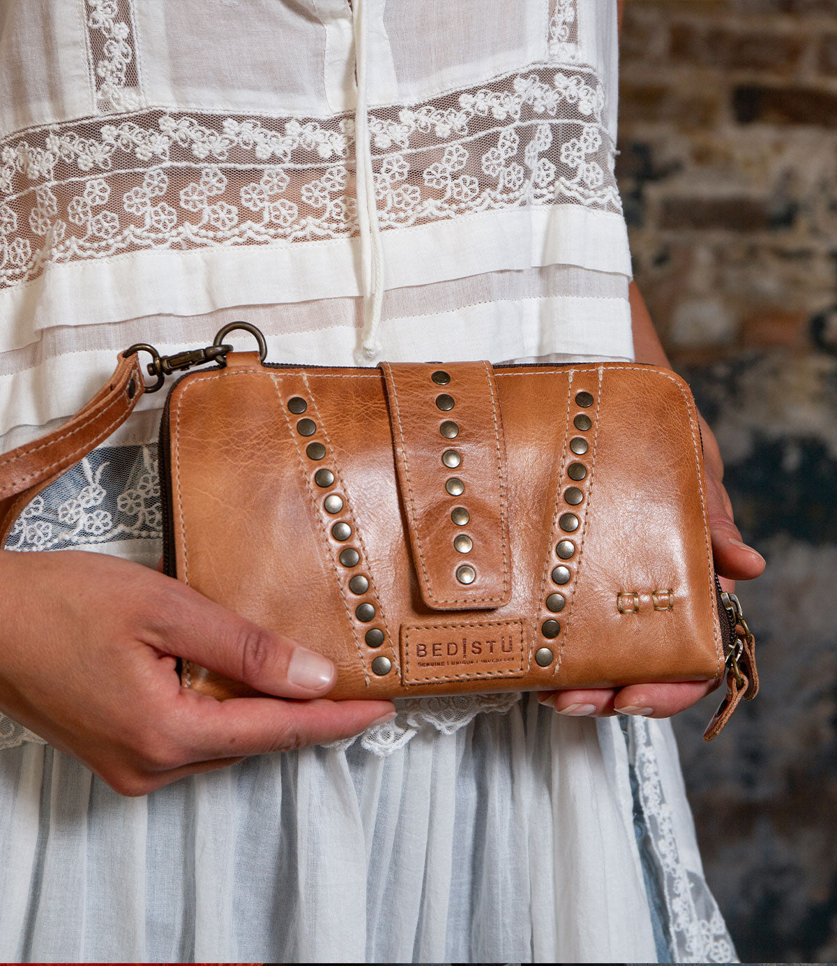 A person in a lace-trimmed white dress holds the Flee clutch by Bed Stu—a brown leather bag with rivet accents, branded label, and a detachable crossbody strap for hands-free use.