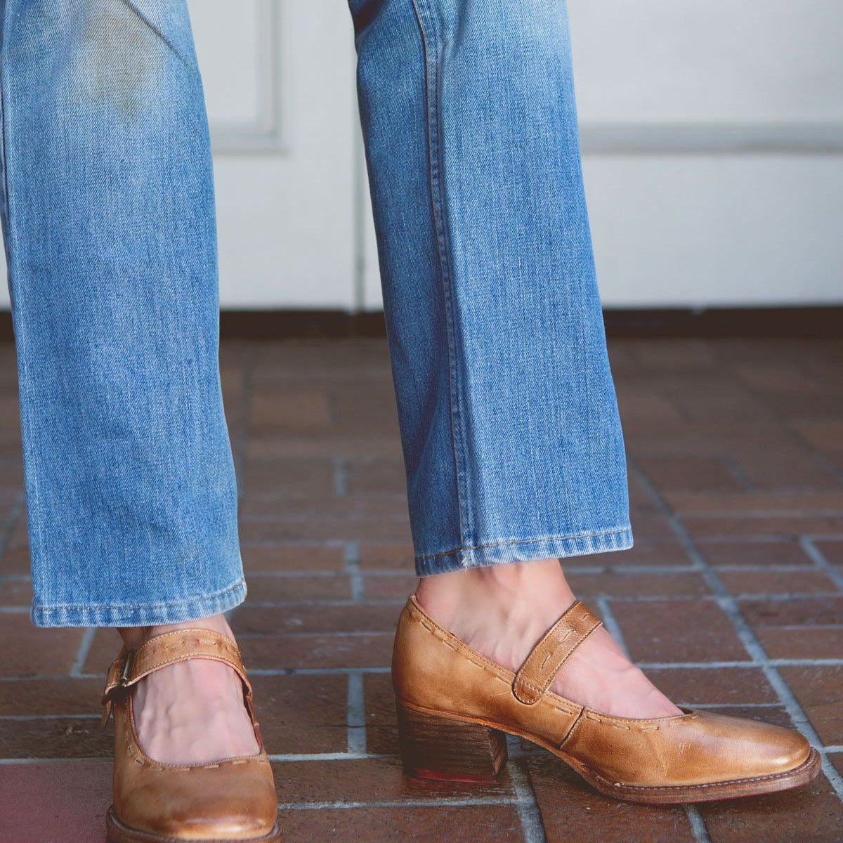 A person wearing light blue jeans and stylish Bed Stu Forefront brown leather Mary Jane shoes with a comfortable heel stands on a brick floor.