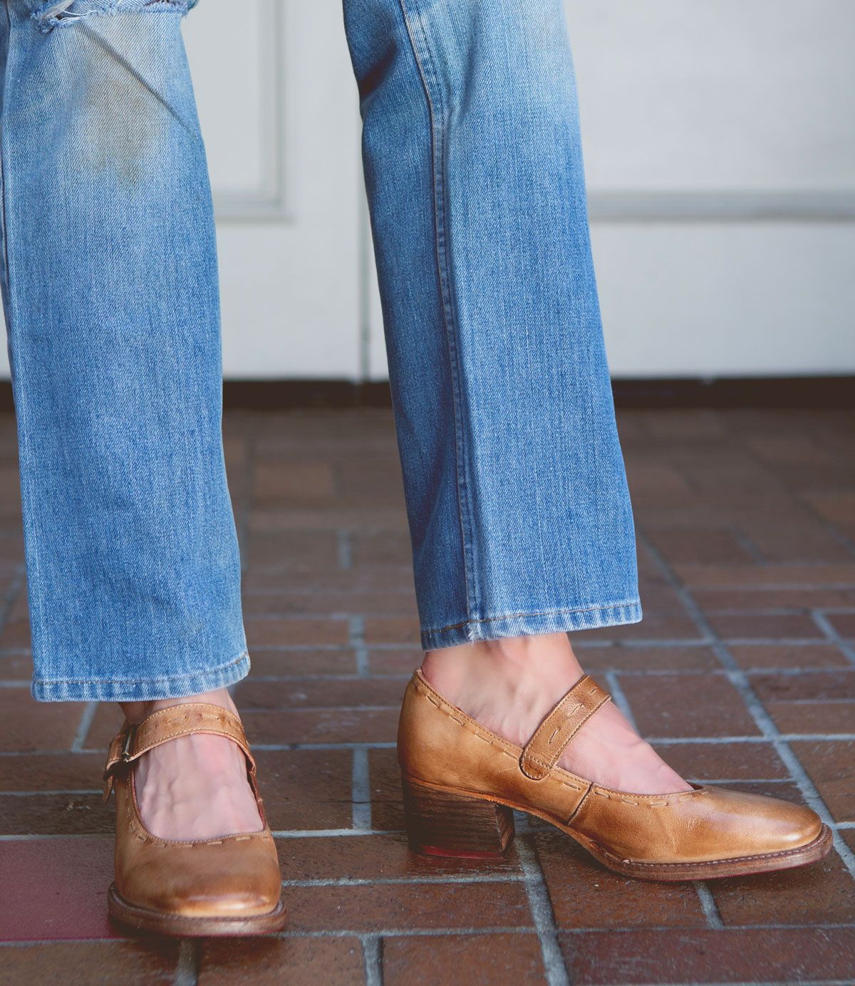 A person wearing light blue jeans and stylish Bed Stu Forefront brown leather Mary Jane shoes with a comfortable heel stands on a brick floor.