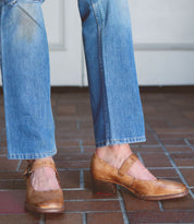 A person wearing light blue jeans and stylish Bed Stu Forefront brown leather Mary Jane shoes with a comfortable heel stands on a brick floor.