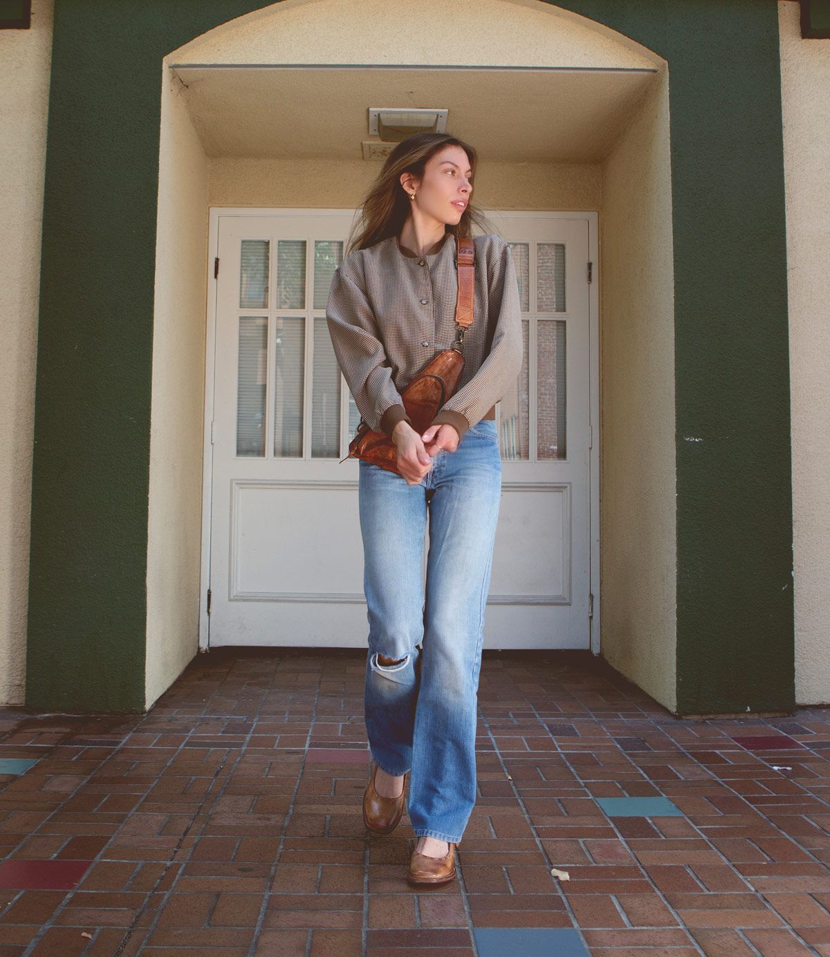 A person stands in front of a door wearing a grey top, blue jeans with a rip on one knee, Bed Stu Forefront brown shoes with an adjustable strap, and a brown bag slung across their shoulder.