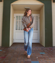 A person stands in front of a door wearing a grey top, blue jeans with a rip on one knee, Bed Stu Forefront brown shoes with an adjustable strap, and a brown bag slung across their shoulder.