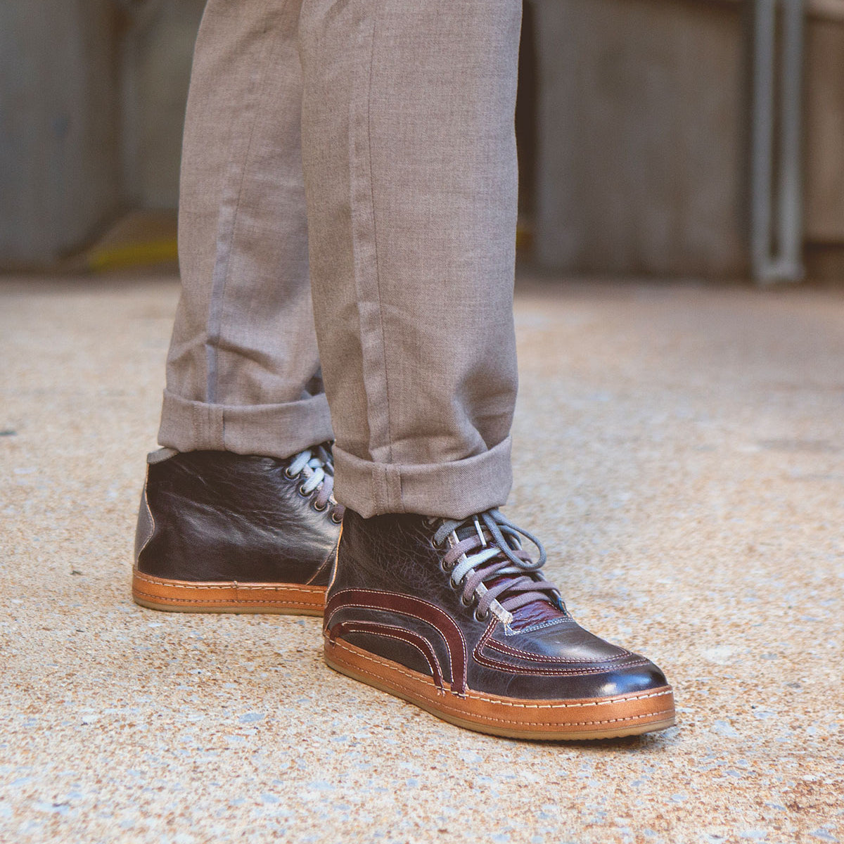A person wearing Bed Stu's Formation brown high-top sneakers featuring subtle leather details, accompanied by beige pants, stands on a textured surface.