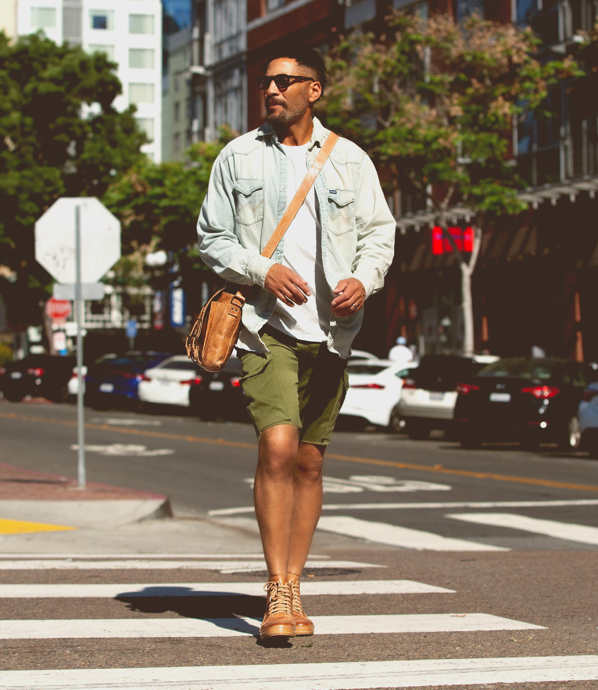A man wearing sunglasses, a light denim jacket, green shorts, and high-top vulcanized sneakers crosses a street at a pedestrian crossing in an urban area. He carries the Formation satchel by Bed Stu.